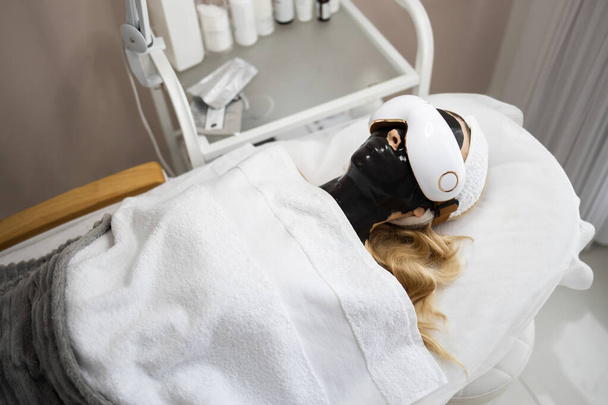 Blonde woman with a black alginate mask on her face and eye massage glasses lies on a couch of a beauty salon. Spa procedures and relaxation	 - Zdjęcie, obraz
