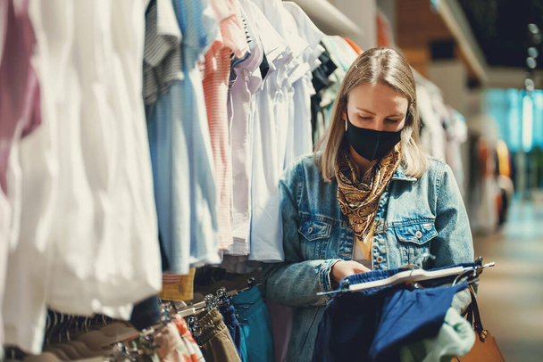 Женщина в медицинской маске выбирает одежду в магазине. - Фото, изображение