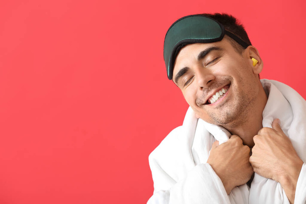 Smiling young man with sleeping mask, blanket and ear plugs on red background - Foto, afbeelding
