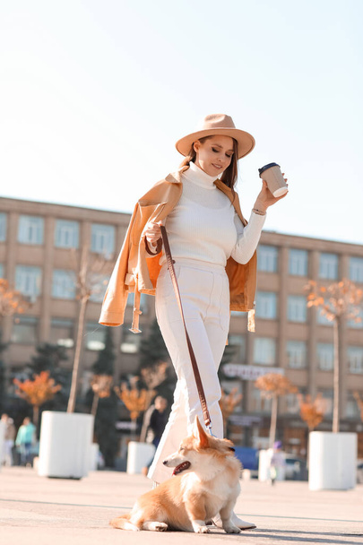 Stylish young woman with cup of coffee and cute Corgi dog walking on city square - Zdjęcie, obraz