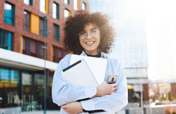 Caucasian woman with curly hair walks on street holds notepad,phone and tablet smiles and looking to camera. Close-up Portrait. Business technology. Curly hair - Foto, immagini