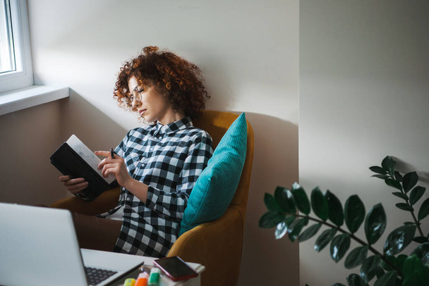 Curly woman in eyeglasses sitting in armchair with laptop on small table aside and taking notes in planner. Freelance career. Business meeting. Business woman - Foto, Imagem