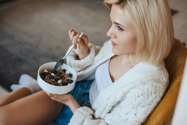 Top view of a bowl of oatmeal with berries in the hands of an caucasian blonde woman. Woman sitting on armachair eating fresh healthy food in the morning - Photo, Image