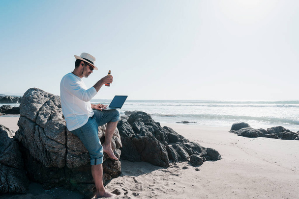 digital nomad κάθεται στην παραλία με ένα φορητό υπολογιστή και πίνοντας μπύρα στο ηλιοβασίλεμα ευτυχισμένη - Φωτογραφία, εικόνα