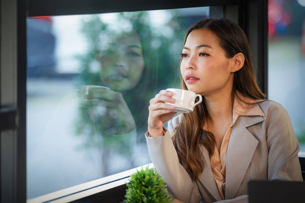 portrait of beautiful asian businesswoman sitting by window in coffee shop holding coffee cup with reflection on window - Fotoğraf, Görsel