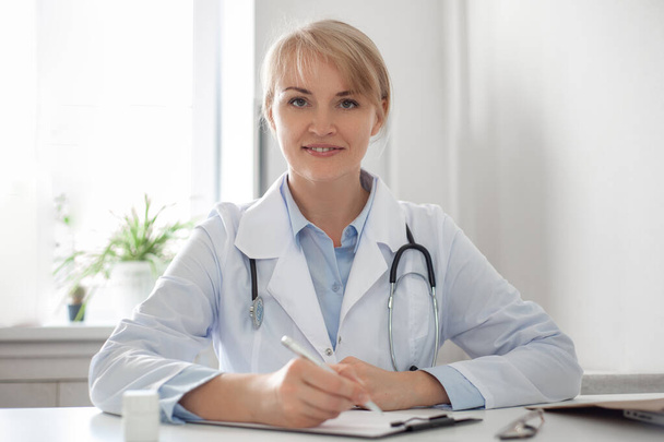 Portrait of a beautiful middle aged female doctor wears white coat stethoscope on shoulders looking at camera. Online counseling and therapy concept Horizontal photo. E-health. Woman smiling. - Foto, Imagem