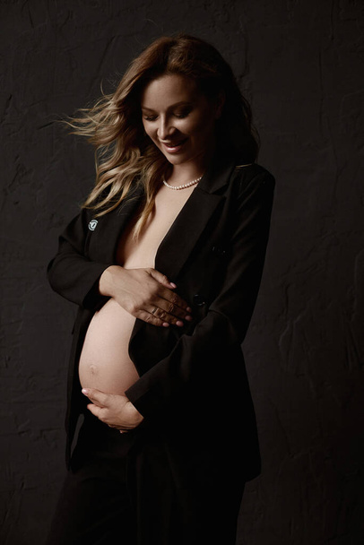 A gorgeous pregnant young woman with fluttering blond hair wearing an unbuttoned blazer embracing her belly and posing beside the black wall. Naked belly. Lifestyle, pregnancy, health - Fotografie, Obrázek