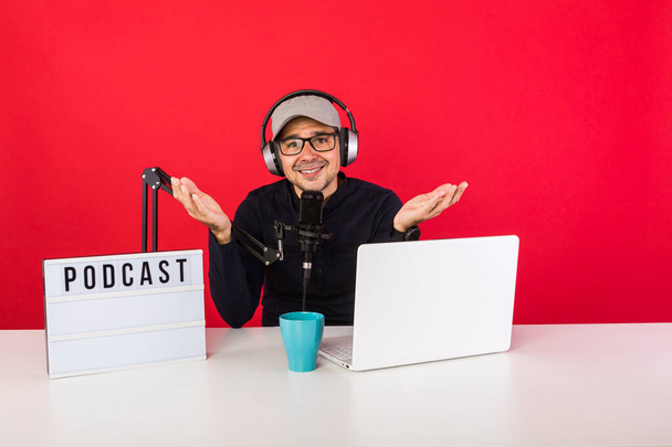 Man presenter with cap in podcast radio recording studio making hand gestures, next to a computer, a microphone and a light box with the word podcast, on red background. Podcasting, broadcast concept - Fotografie, Obrázek