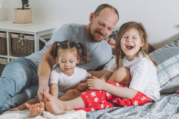 Father plays with two children on bed. Dad tickles kids feet. Family of daddy, and two little girls - Fotografie, Obrázek