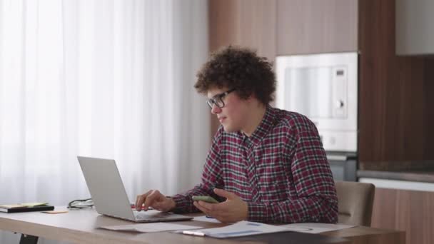 Young man with smartphone in his hands. Modern businessman or student at home office. Freelancer at work. Young student man study at home with laptop and uses a smartphone - Footage, Video