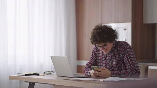 Young man with smartphone in his hands. Modern businessman or student at home office. Freelancer at work. Young student man study at home with laptop and uses a smartphone - Materiał filmowy, wideo
