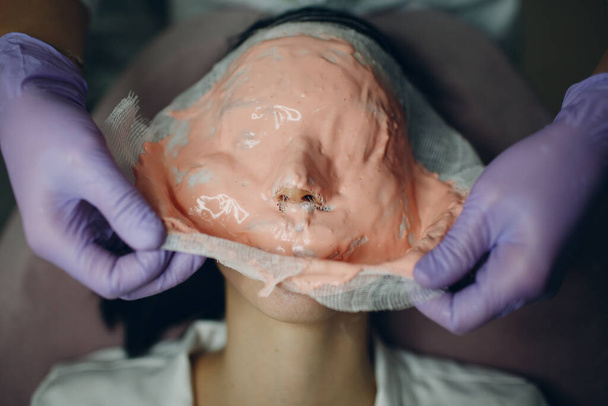 Alginar o processo de remoção da máscara facial. As mãos femininas de um esteticista segurando espátula. Mulher deitada em um sofá no escritório do cosmetologista. Cuidados com a pele facial no salão de beleza spa - Foto, Imagem