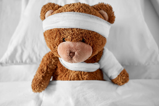 ill teddy bear with bandaged paw and head in bed - Valokuva, kuva