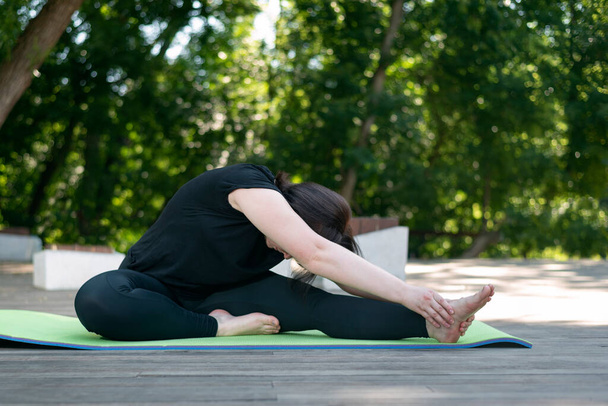 Yoga class in the morning in the park. Side view. Young beautiful flexible girl makes a lean forward with her hands from a sitting position - Foto, Imagem