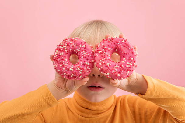Fair-haired baby holds pink donuts near his eyes. Portrait of boy with doughnut on pink background - Photo, Image