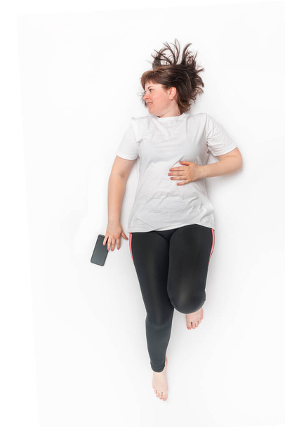 Portrait of plus size woman in sportswear lies and looks away on white background. Model with curvy figure. Top view. - Zdjęcie, obraz