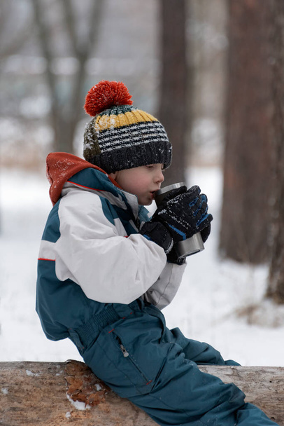 Child in the winter forest drinks hot tea from a thermomug. Portrait of boy in knitted hat in snowy forest - Fotó, kép