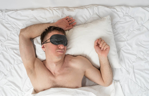 Young man lies in bed in sleep mask under the white sheet. Portrait of sleeping man top view - Fotoğraf, Görsel