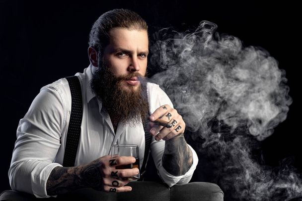 Portrait of hipster brutal bearded man with cigar and a glass whiskey in a dark room. Tattoos hands - Foto, Imagem