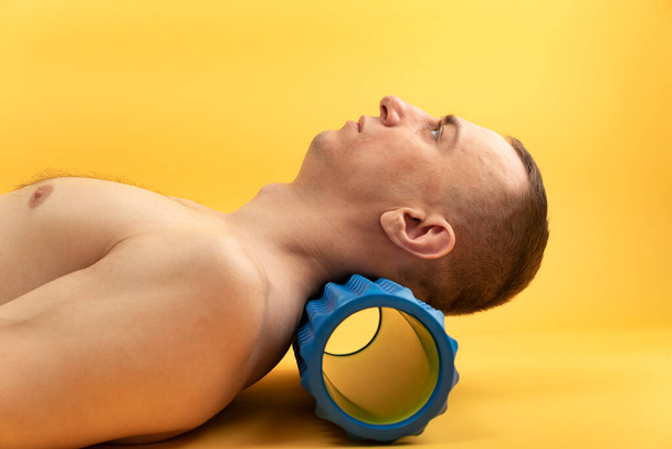 Άνδρας αθλητής ανακουφίσει πονόλαιμο μύες μετά την προπόνηση σε κίτρινο φόντο. Ο άντρας ξαπλώνει στην πλάτη με ένα ρολό μασάζ κάτω από το λαιμό του. - Φωτογραφία, εικόνα
