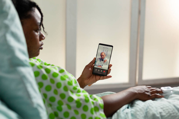 Paciente afroamericano enfermo sosteniendo el teléfono discutiendo la experiencia de la enfermedad con el médico terapeuta remoto - Foto, Imagen