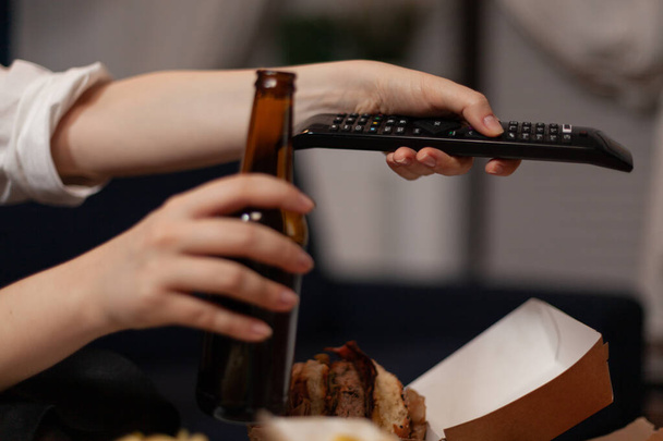 Κοντινό πλάνο των χεριών κρατώντας μπουκάλι μπύρα χαλαρωτικό περιήγηση τηλεοπτικά κανάλια χρησιμοποιώντας τηλεχειριστήριο - Φωτογραφία, εικόνα