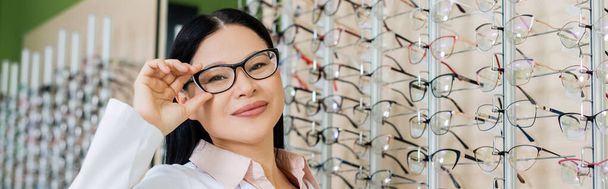 happy asian oculist looking at camera and touching eyeglasses in optics salon, banner - Фото, изображение