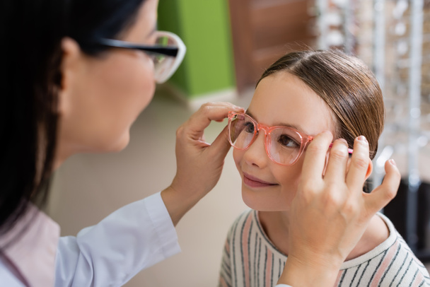 blurred oculist trying eyeglasses on smiling girl in optics shop - Foto, Imagem