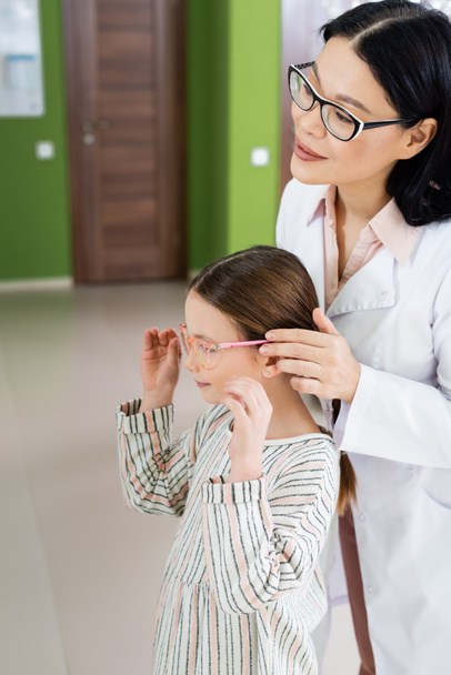 positive asian ophthalmologist in white coat trying eyeglasses on child in optics salon - Fotó, kép