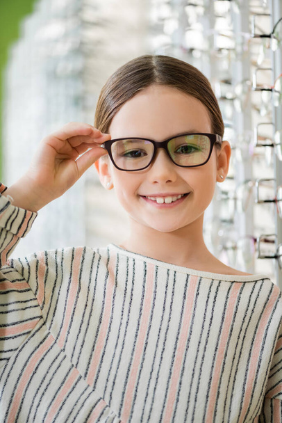 pleased child looking at camera while trying on eyeglasses in optics shop on blurred background - Photo, Image