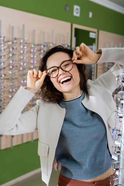 excited woman laughing at camera while trying on eyeglasses in optics shop - Foto, Bild