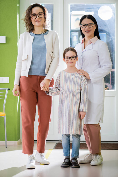mother and daughter with asian ophthalmologist in eyeglasses smiling at camera in optics shop - Фото, изображение