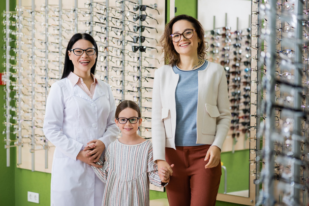 happy girl in eyeglasses holding hands with mom and asian oculist while looking at camera in optics salon - Foto, Bild