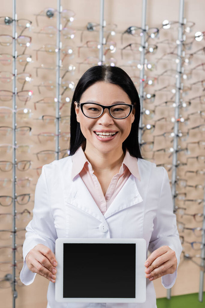 happy asian oculist in eyeglasses holding digital tablet with blank screen in optics shop - Foto, Bild