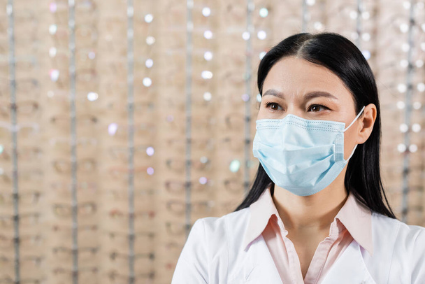 азіатський окуліст в медичній масці біля розмитого асортименту окулярів в салоні оптики
 - Фото, зображення