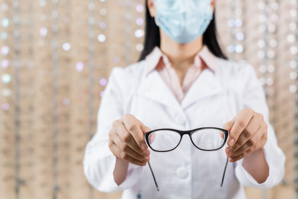 cropped view of ophthalmologist in medical mask showing eyeglasses in optics store - Foto, immagini