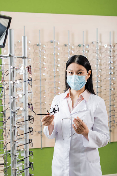 asian oculist in medical mask comparing eyeglasses while working in optics shop - Foto, afbeelding