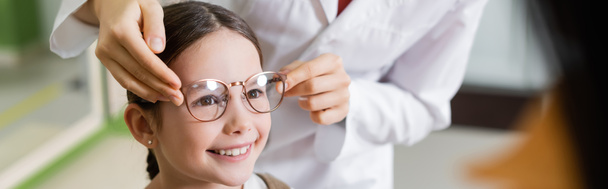 oculist trying glasses on smiling girl near blurred mother in optics store, banner - Фото, изображение