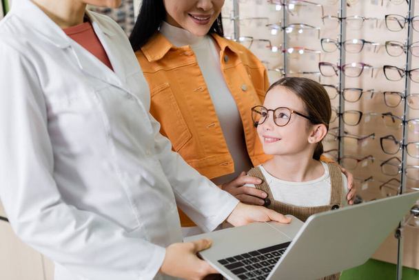pleased child in eyeglasses smiling near mother and oculist with laptop in optics shop - Foto, afbeelding