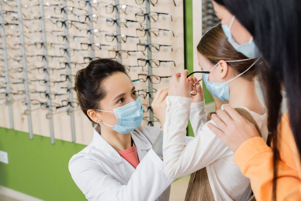 oculist in medical mask trying glasses on girl near blurred asian woman in optics store - Foto, immagini