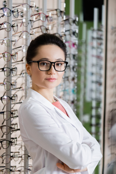 oculist in white coat standing with crossed arms near assortment of eyeglasses in optics shop - Foto, Imagem