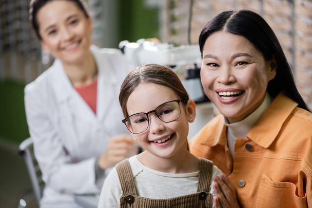 happy kid in eyeglasses looking at camera near asian mom and blurred ophthalmologist in optics shop - Фото, изображение