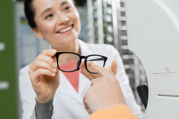 woman pointing with finger at eyeglasses in hand of happy oculist in optics shop - Foto, Imagem