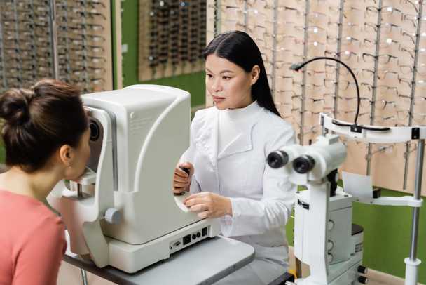 asian optometrist checking vision of blurred woman on ophthalmoscope in optics store - Foto, afbeelding