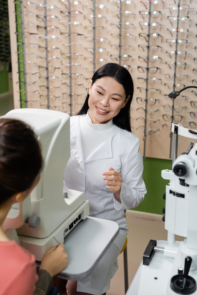 sonriente asiático oculista prueba visión de borrosa mujer en oftalmoscopio en óptica tienda - Foto, Imagen
