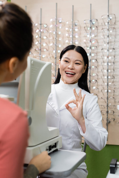 ευχαριστημένη ασιατική οφθαλμίατρος δείχνει εντάξει χειρονομία κοντά screener όρασης και θολή γυναίκα στο σαλόνι οπτικών - Φωτογραφία, εικόνα