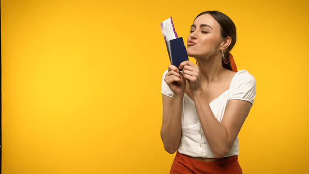 Νεαρή γυναίκα φιλάει διαβατήριο και αεροπορικό εισιτήριο απομονωμένο στο κίτρινο - Φωτογραφία, εικόνα