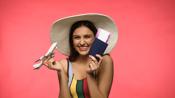 Happy woman in swimsuit holding toy plane and passport isolated on pink  - Foto, Imagen