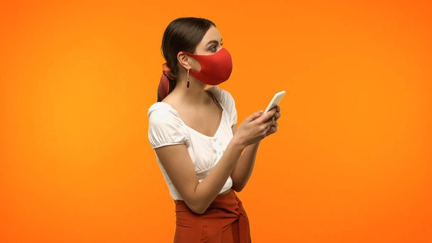 Brunette woman in protective mask holding smartphone isolated on orange  - Foto, Imagem