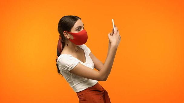 Young woman in protective mask taking photo on smartphone isolated on orange  - Φωτογραφία, εικόνα